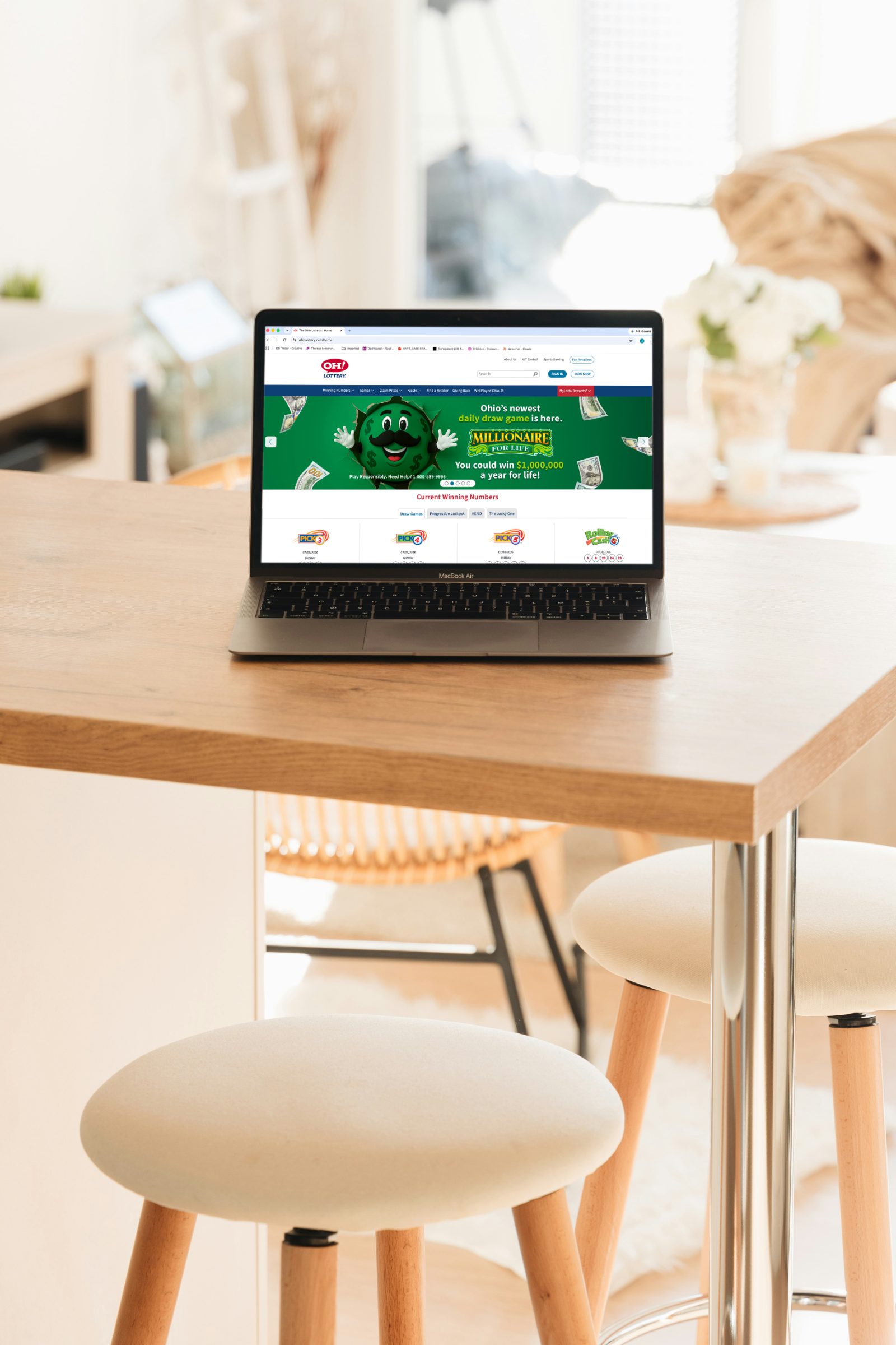 Laptop on a light wood kitchen table showing the OH! Lottery homepage with a Millionaire For Life hero banner announcing Ohio's newest daily draw game.