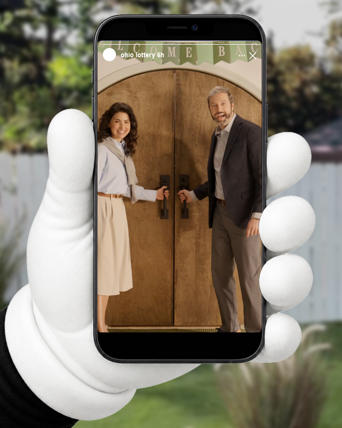 A white-gloved hand holding a phone in a backyard, showing an Ohio Lottery Instagram Story of a smiling couple opening wooden double doors beneath a Welcome Back banner.